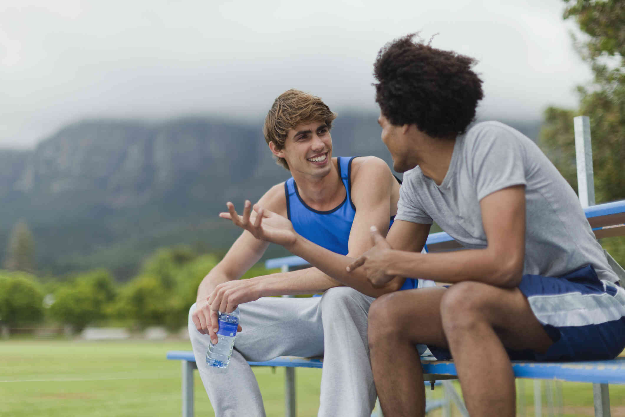 Two men in workout clothes sit side by side on outdoor bleachers while talking and smiling.